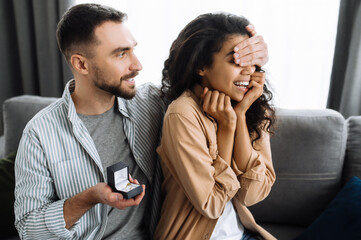 Multiracial couple in love at home on the living room sofa. The guy closes the eyes of his beloved girlfriend with his hand, preparing to give her a ring and make make a marriage proposal.