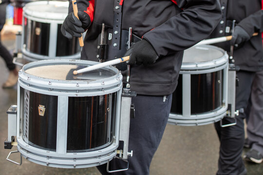 A young man wearing dark clothing holds two drumsticks and beats on a black and white drum at an outdoor concert or parade. There are multiple drummers performing in a music band with a percussionist.