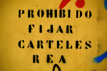 Spanish text forbidding the posting of notices or posters spray painted onto a brown wall. This is...