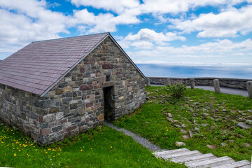 An old one level building made from multi coloured rocks and a pink slate roof. There's a bright blue sky with white clouds in the background, vibrant green grass in the foreground and wooden steps.