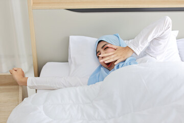 Beautiful asian muslim woman wearing white sleepwear lying on bed, stretching her arms after getting up in the morning at sunrise. Cute young girl with hijab wake up and relaxing, closing her eyes