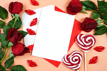 Happy Valentine's Day greeting card and envelope, styled with Valentine red roses, heart shape gift and lipstick kissess chocolates on a modern coral background. Mockup. Top view flat lay. Copy space.