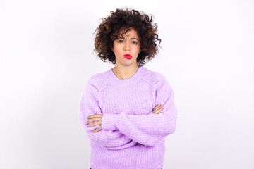 Gloomy dissatisfied young beautiful caucasian woman wearing pink knitted sweater against white wall looks with miserable expression at camera from under forehead, makes unhappy grimace