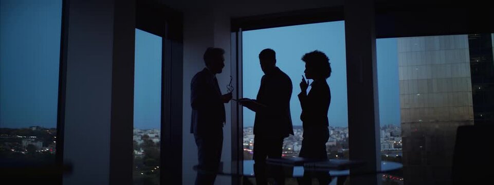 Business people discussing in late evening in office. Blue hour light scene. Overtime hours. Silhouettes against large window.