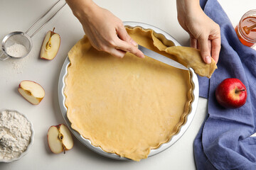 Woman cutting dough leftovers for traditional English apple pie in baking dish at white table, top...