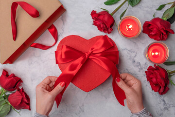 Women's hands touch a red satin bow on a cardboard heart-shaped gift box. Lit candles, roses, craft shopping bag, white marble. Valentine's Day 14 february, Engagement, wedding anniversary