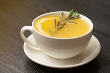 Yellow ginger tea with lemon and rosemary branch, in white cup on wooden table