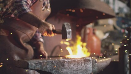 Close up of male blacksmith hammering metalwork on anvil with sparks and blazing forge in background - shot in slow motion - Powered by Adobe