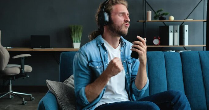 Slow motion of handsome Bearded Hipster guy have Fun jumping dancing on bed and listening to Music in Headphones. Man playing imaginary Guitar in Loft Living Room. Playfully Mood at Home. Funny Man.