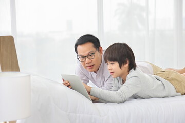 Asian father teaching online homework to his son in bed for homeschooling and education concept