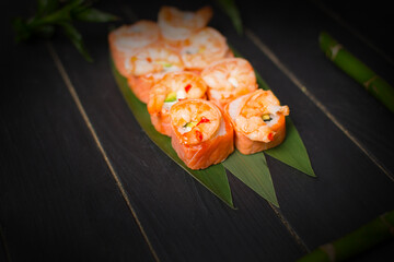 Tasty Japanese seafood sushi roll with salmon tataki and roasted black tiger shrimps on top served on bamboo leaves with bamboo plant and sticks on black wooden background
