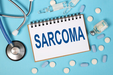 SARCOMA, text on white notepad over blue background. blue stethoscope. pills. ampoules