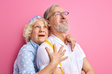elderly woman stand in shock with opened mouth hugging man from back, they look up, isolated on pink background