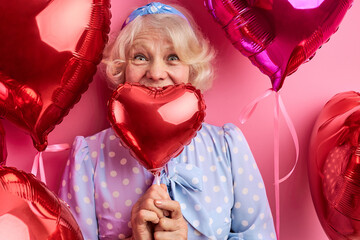 gray-haired woman with heart shaped air balloons isolated on pink background, happy woman smiles. dreamy woman in party dress celebrating