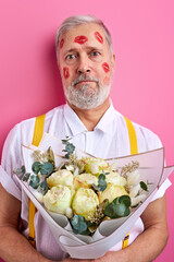 senior man with a bouquet of flowers waiting on a date, with lipstick marks on face. isolated on pink background