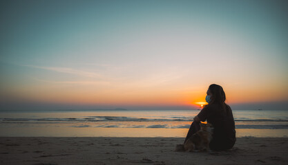 Fototapeta premium Woman wearing surgical mask for prevent coronavirus Outbreak with small dog travel on the beach at sunset time in Thailand.