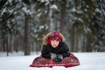 Fototapeta na wymiar A smiling teenage boy in a black jacket with a fur and red hat lies on a tubing in a snow-covered forest. Winter leisure.