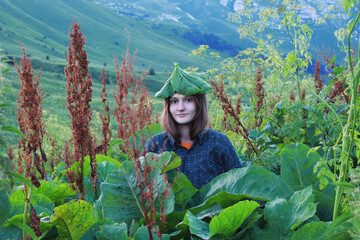 A young beautiful woman stands in a field among large plants. On the girl's head is a hat made of burdock leaf. Eco-friendly life concept. Life in nature
