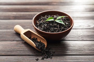 Dry black tea leaves in bowl and scoop on wooden background