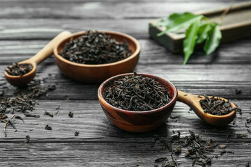 Dry black tea leaves in bowls and spoons on wooden background