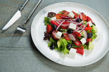Macro shot of Traditional Greek salad on marble plate served with fork, knife. Salad with Feta cheese, black olives, lettuce, fresh cucumber, cherry tomatoes served with onion on stone background
