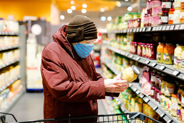 Old senior woman wear medical mask, protection against pandemic coronavirus disease. 90 years retired female lady push cart trolley for shopping in supermarket. Woman buy food and groceries