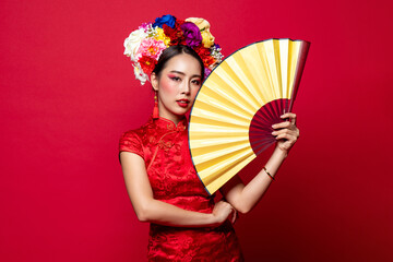 Obraz premium Beautiful Asian woman wearing traditional cheongsam qipao dress with colorful flowers on head holding golden fan in isolated studio red background