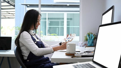 Side view of young female artist or designer painting with water color while sitting at workspace.