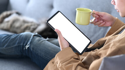 Close up view of young man using digital tablet and holding a cup of coffee while resting on sofa.