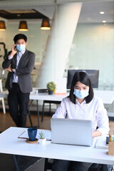 Portrait of businesswoman wearing protective mask working on laptop and her colleague talking on mobile phone in background.