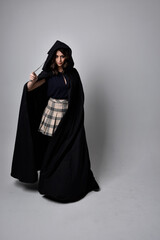 full length portrait of pretty brunette woman wearing tartan skirt and boots with long black cloak.. Standing pose holding a magical wand against a  studio background.