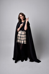 full length portrait of pretty brunette woman wearing tartan skirt and boots with long black cloak.. Standing pose holding a magical wand against a  studio background.