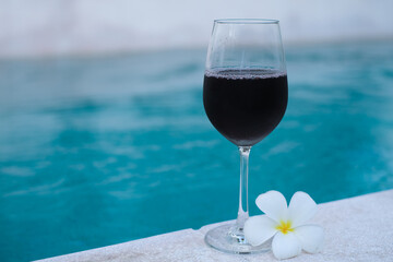 Glass of red wine and white frangipani tropical near a swimming pool in morning of summer. 
