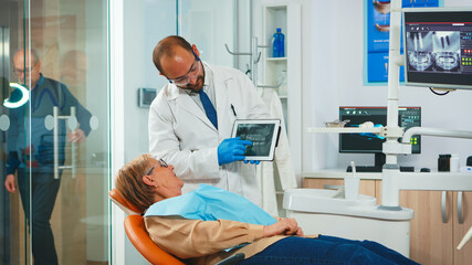 Stomatologist reviewing x ray of tooth with senior patient explaining treatment. Dentist showing to old woman dental radiography using tablet, doctor and nurse working together in modern clinic.