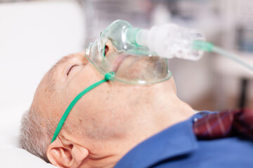 Senior man doing inhalation through oxygen mask because of covid19. Patient in hospital room laying...
