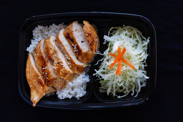 Grilled teriyaki chicken with rice in a lunch box on black background.
