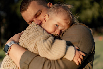 young dad with eyes closed hugging baby son putting head to his shoulder and sucking a pacifier. they are standing in park