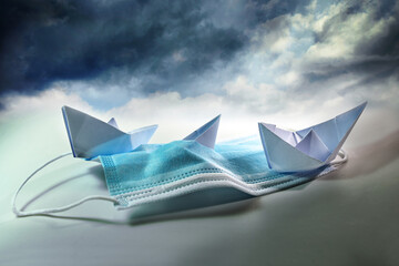 Three paper boats struggle against sinking on a surgical face mask under a stormy sky, metaphor for...