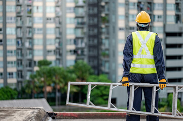 Asian maintenance worker man with safety helmet and reflective suit carrying aluminium step ladder...
