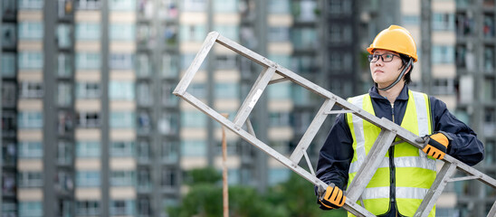 Asian maintenance worker man with safety helmet and reflective suit carrying aluminium step ladder...