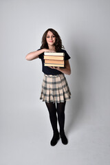 full length portrait of pretty brunette woman wearing tartan skirt and boots.  Standing pose holding books against a  studio background.