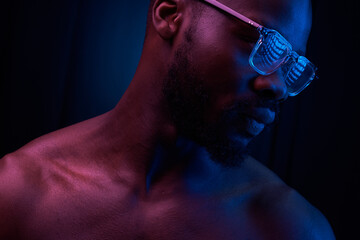 portrait of a handsome black man in red and blue light on dark background  with glasses on his eyes, he has naked sports torso