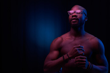 portrait of a handsome black man in red and blue light on dark background  with glasses on his eyes, he has naked sports torso and bracelets on arms