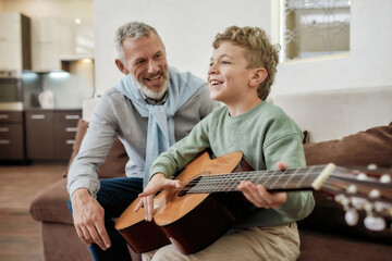 Excited little boy learning how to play acoustic guitar with grandfather, they sitting together on...