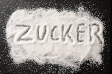 German word Zucker, meaning sugar, written in spilled out sugar crystals on a dark slate, high...