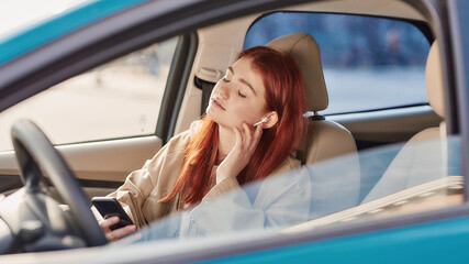 Teenage girl in earphones sitting in the car with her eyes closed while using her phone, listening to music