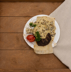 Lamb steak lies on a white plate, on a thin pita bread. Top with mushrooms in a creamy sauce. Decorated with chopped tomatoes, curly parsley sprigs. The background is brown boards, a beige napkin.