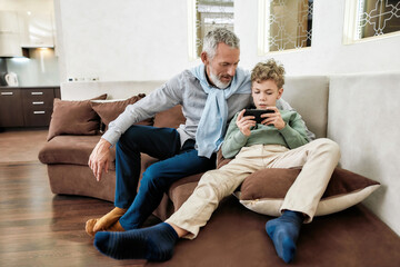 Cute little boy grandson watching video or playing video game on smartphone while sitting on sofa with his grandfather, spending time together at home