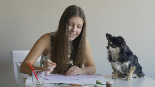 Teen age girl painting by the table with her doggy, smiling beauty and a pet.Chihuahua resting.Art activity during lockdown,pleasure of having pet as a friend. Cute domestic picture, a day in a life.
