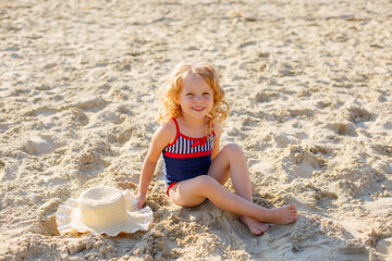 cute little curly-haired girl in a swimsuit sitting on the beach on the sand in summer on vacation